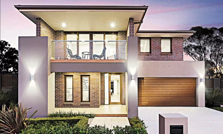 A modern two-story house with brick and neutral tones, large windows, a balcony with outdoor seating, and a double garage with a wooden door, surrounded by landscaped greenery at dusk.