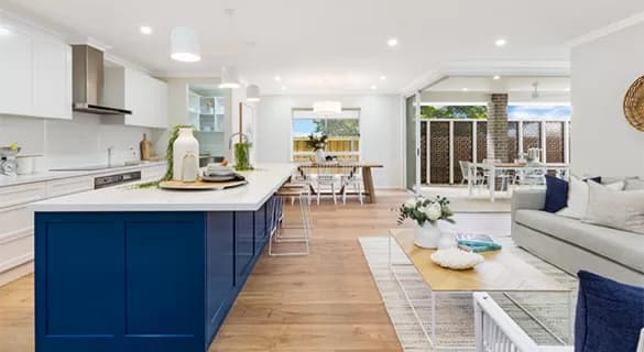 Modern open-plan living space featuring a kitchen with a blue island, white cabinets, dining area, and a cozy living room with a gray sofa and coffee table, leading to an outdoor patio.