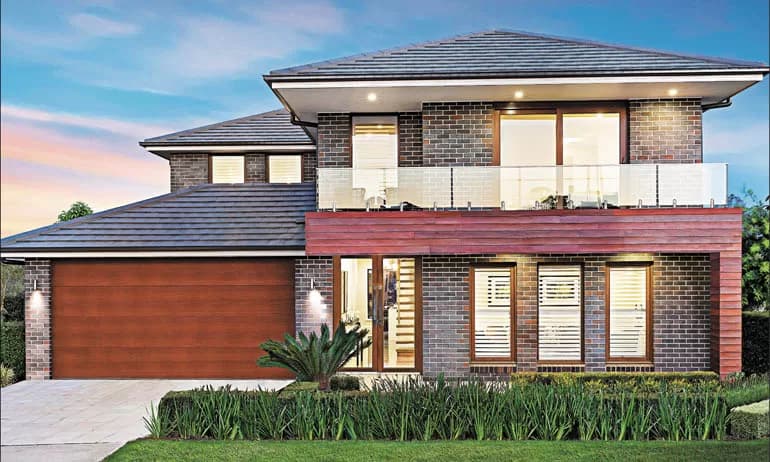 Modern two-story house with dark brick exterior, large windows with white shutters, a wooden double garage door, glass balcony, well-kept lawn, and shrubs in the front garden at sunset.