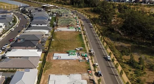 Aerial view of a suburban neighborhood showing houses, vacant lots, and several homes under construction along a curved road lined with trees. Construction materials and vehicles are visible on some of the lots.