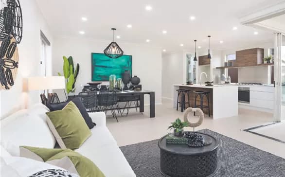 Modern open-plan living area with a white sofa, green and black cushions, a black round coffee table, dining table with chairs, wall art, kitchen island with stools, and contemporary decor in bright, natural light.