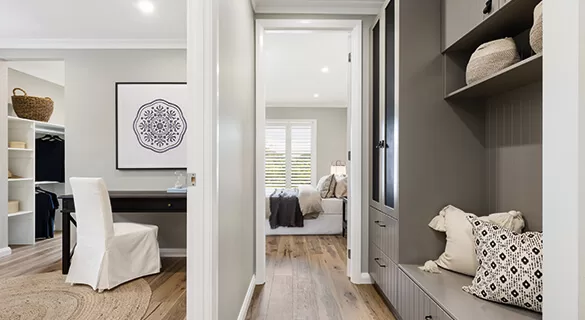 A hallway with built-in gray cabinets, open shelves, and decorative baskets leads to a bedroom with a neatly made bed. A home office with a white chair and desk is visible on the left. Light wood floors run throughout.