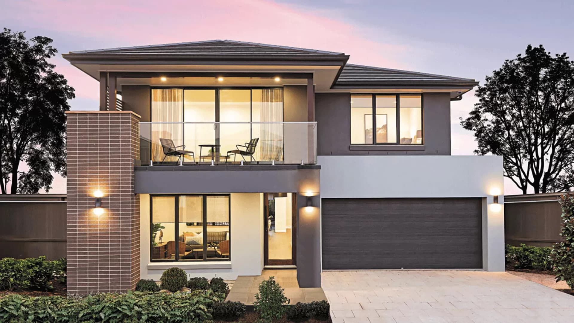 Modern two-story house with large windows, a balcony with chairs, brick and gray accents, double garage, and soft outdoor lighting at sunset, surrounded by small shrubs and trees.