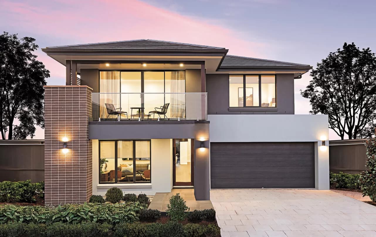 Modern two-story house with large windows, a balcony with two chairs, brick and gray exterior walls, and a double garage. The driveway is surrounded by neatly trimmed bushes and plants; evening sky in the background.