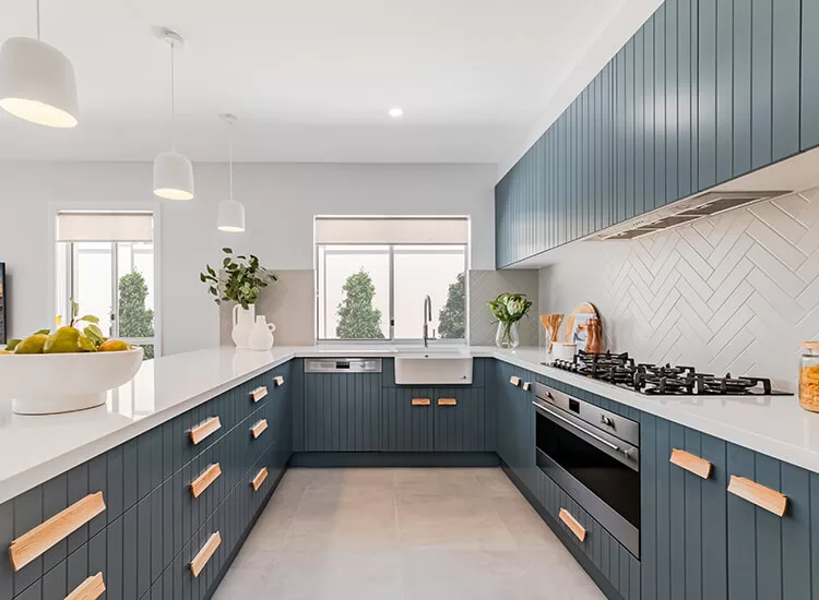 Modern kitchen with blue cabinets, wooden handles, white countertops, built-in oven, gas stove, and herringbone tile backsplash. Large windows provide natural light; pendant lights hang above the counter.