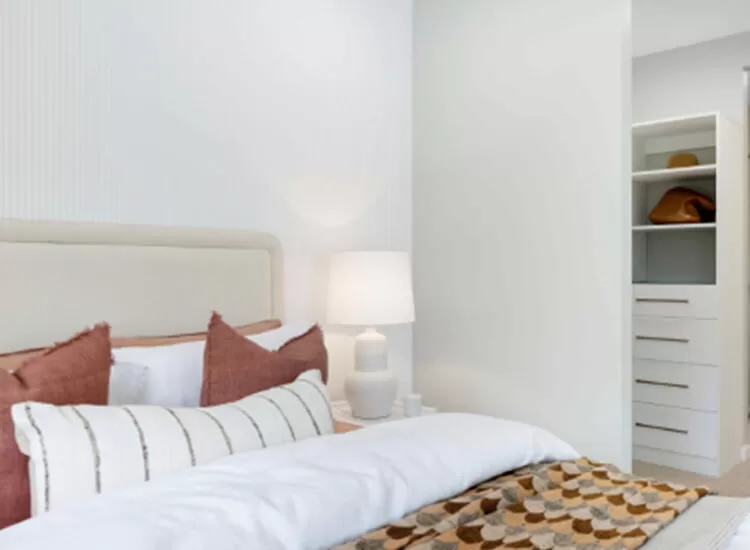 A cozy bedroom with a white headboard, pink and white pillows, a white lamp on a nightstand, and a patterned blanket. An open closet with shelves and drawers is visible in the background.
