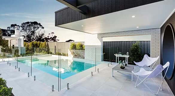 Modern backyard with a swimming pool enclosed by a glass fence, white lounge chairs, small table, potted plants, and surrounding greenery under a partially covered patio.
