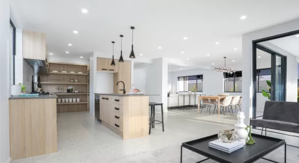 Modern open-plan kitchen and dining area with light wood cabinetry, a central island, black pendant lights, and a dining table in the background. A small coffee table and armchair are in the foreground. The space is bright and airy.