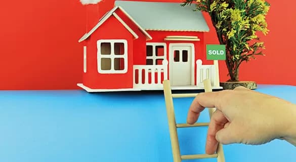 A hand holds a small wooden ladder leading up to a red toy house with a SOLD sign, set against a blue and red background with a model tree beside the house.