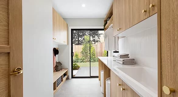 Modern laundry room with light wood cabinets, white countertops, and a sink. Stacked folded towels sit by the sink. Glass sliding doors open to a green garden with shrubs and a wooden fence outside.