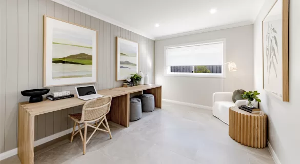 Modern home office with a long wooden desk, laptop, and rattan chair. Two landscape paintings are on the wall. Theres a white armchair, round side table with a plant, and soft natural lighting from a window.
