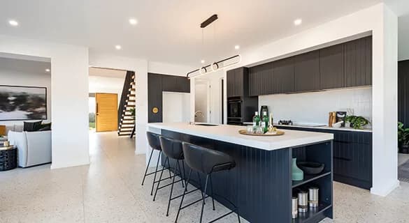 Modern kitchen with black cabinets, a white countertop island with two black barstools, pendant lighting, and sleek decor. Open layout leads to a living area and a wooden staircase with a glass railing.