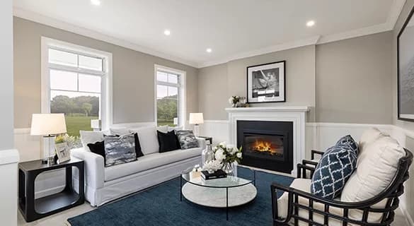A modern living room with a white sofa, a patterned armchair, a glass coffee table with flowers, a blue rug, and a fireplace beneath a framed photo. Large windows provide natural light and a view of greenery.