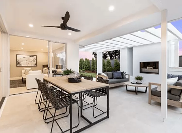 Modern covered patio with a wooden dining table and black chairs, ceiling fan, lounge seating, outdoor fireplace, and sliding glass doors leading to a bright living room. Greenery and trees visible outside.