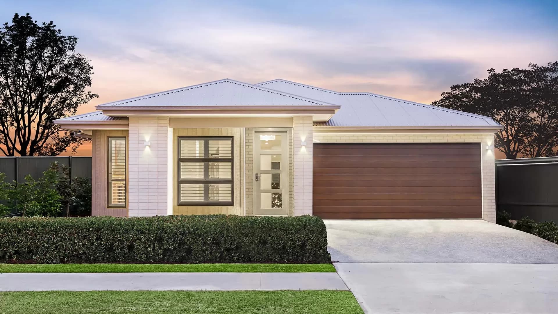 Modern single-story house with light-colored brick exterior, large windows, frosted glass front door, and a double wooden garage. Neatly trimmed bushes and lawn in front, with trees and a pastel sunset sky in the background.