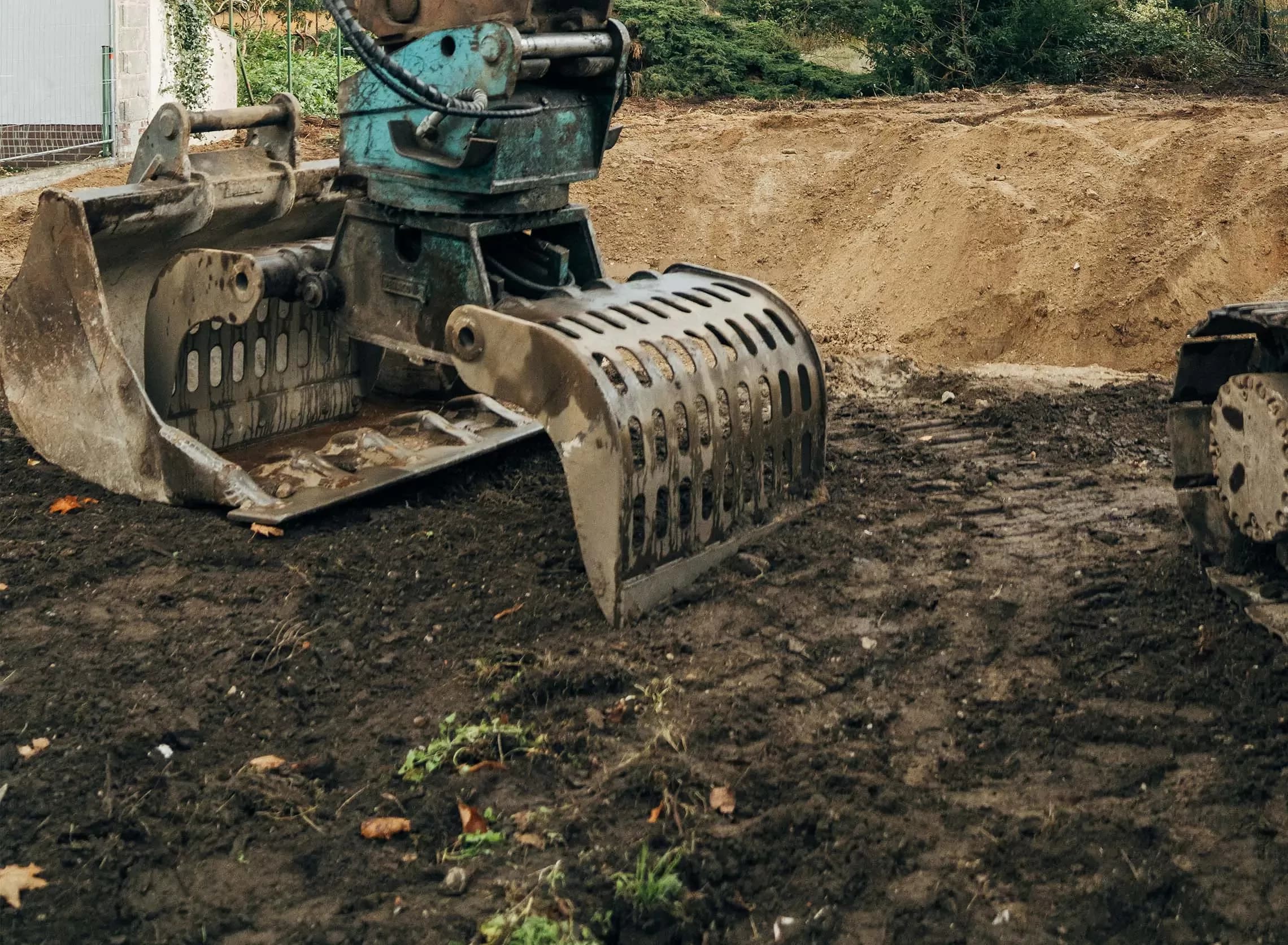 An excavator with a metal grapple attachment sits on a dirt construction site, with a mound of soil and some green plants visible in the background.