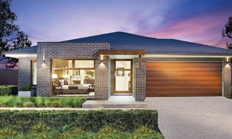 Modern single-story brick house at dusk with large front window showing a lit living room, wooden double garage door, landscaped front yard, and pathway leading to the main entrance.