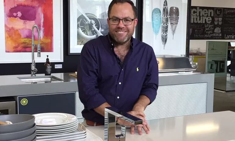 A smiling man in glasses and a dark shirt stands in a modern kitchen, washing his hands at a faucet mounted on a countertop next to a stack of plates, with colorful artwork and chalkboard menus in the background.