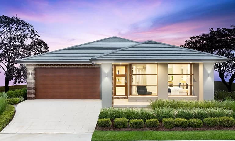 Modern single-story house with a large wooden garage door, wide windows showing a well-lit interior, a neatly trimmed front lawn, and a paved driveway. The sky is colorful with purple and blue hues at dusk.
