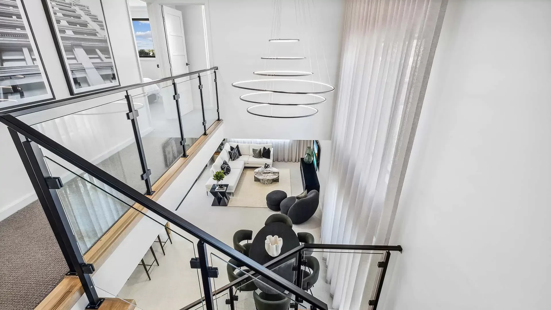 Modern, bright living room viewed from a glass-railed staircase; features a circular chandelier, sleek black and gray furniture, white walls, large windows with sheer curtains, and abstract artwork.