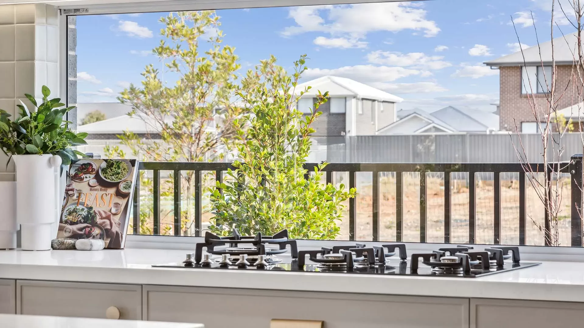 A modern kitchen with a gas stove on a white countertop, a cookbook on display, and a large open window overlooking a backyard with trees, a fence, and neighboring houses under a blue sky.