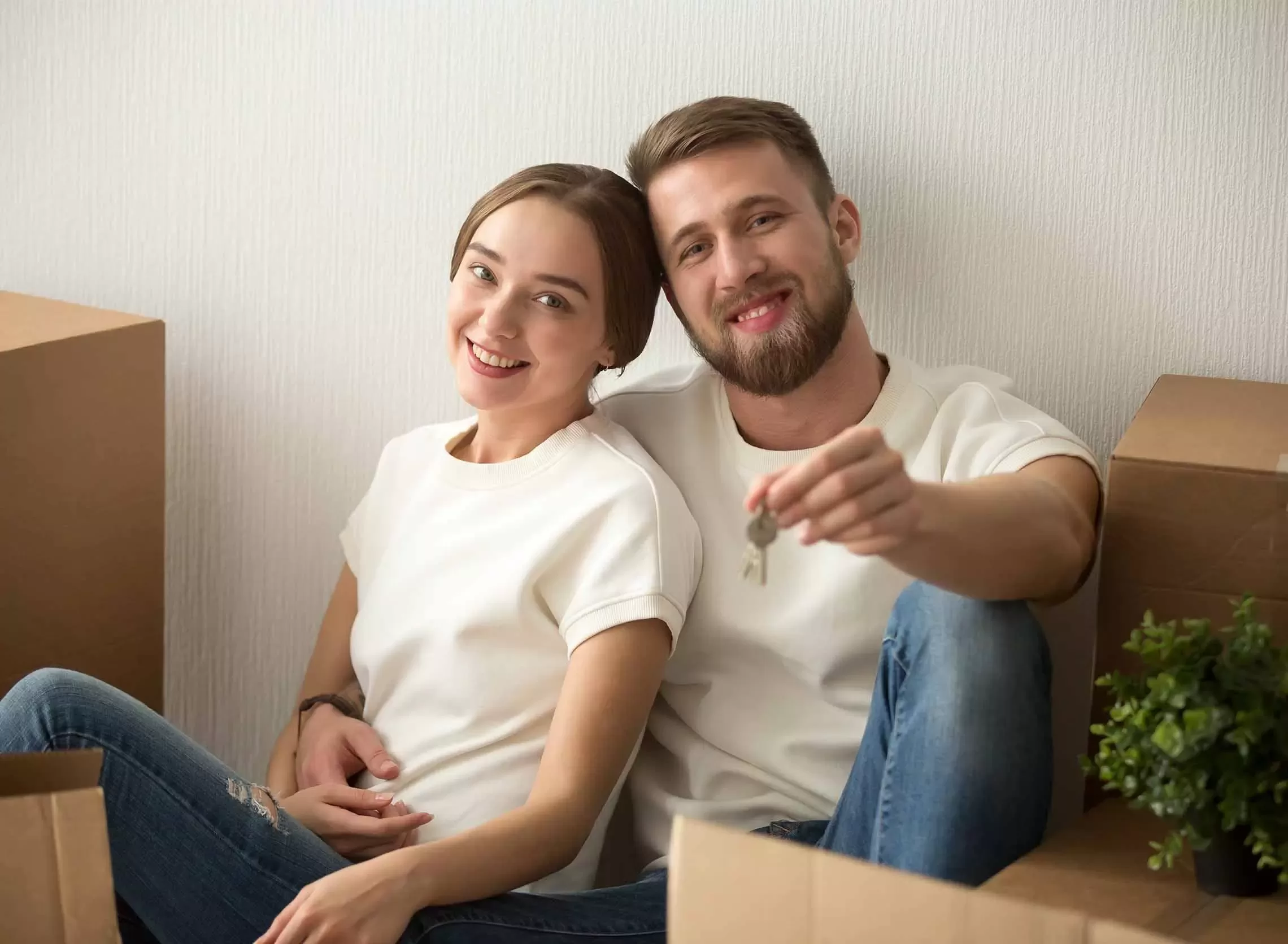 A smiling couple sits on the floor among moving boxes. The man is holding a key, and they both look happy and relaxed, suggesting they have just moved into a new home. A small potted plant is nearby.