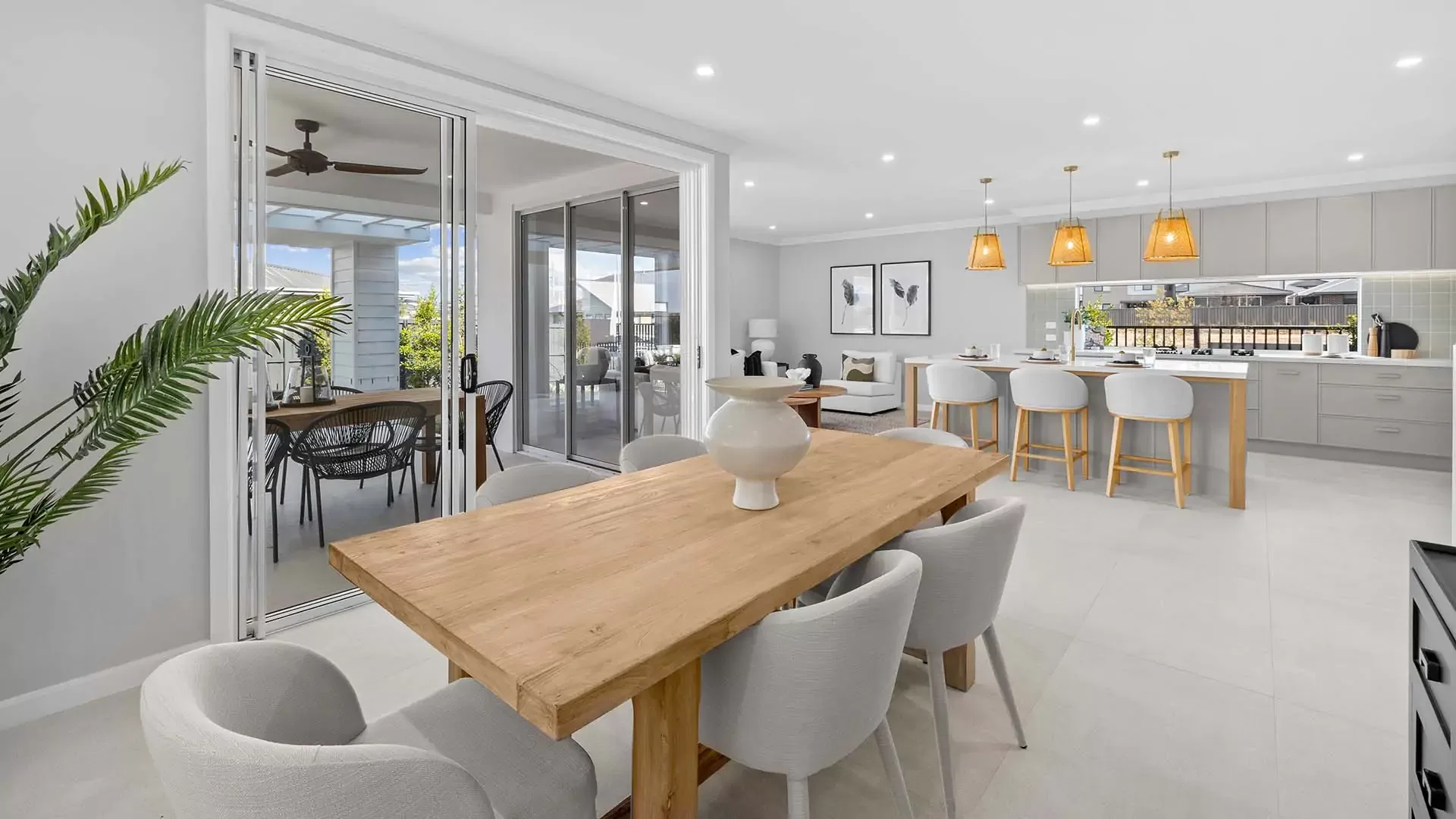 Modern open-plan dining and kitchen area with light wood furniture, white chairs, pendant lights, and sliding glass doors leading to an outdoor patio with additional seating. Bright, airy, and contemporary decor.