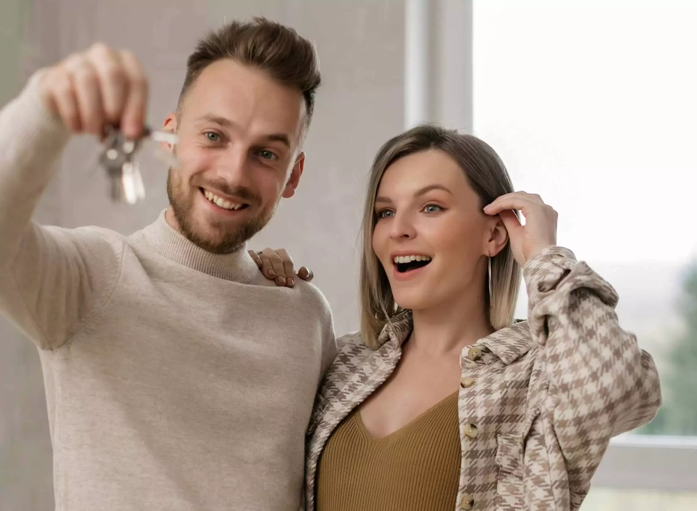A smiling man holds up a set of keys while a woman beside him looks surprised and happy. They stand close together indoors, appearing excited, possibly about a new home.
