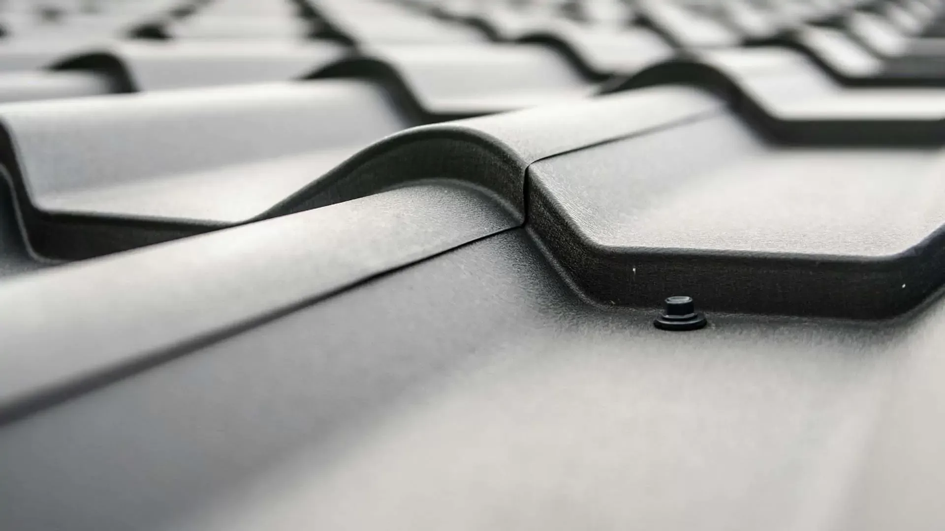 Close-up view of a gray metal roof with wavy, overlapping panels and a visible fastening screw, creating a pattern of ridges and shadows along the surface.