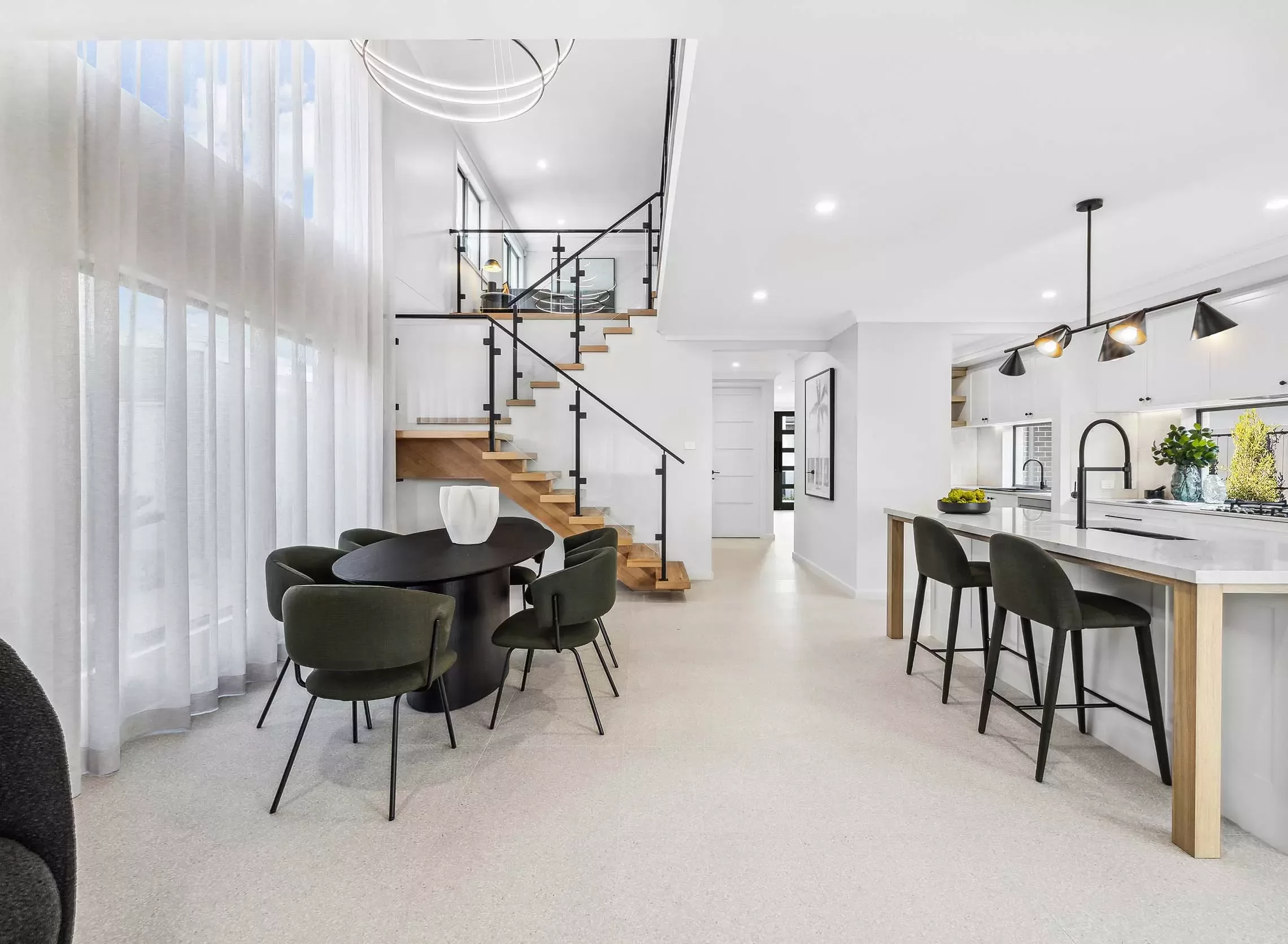 Modern open-plan interior with a kitchen, island with barstools, dining area with round table and dark chairs, wooden staircase with glass railing, large windows with sheer curtains, and minimalist decor.