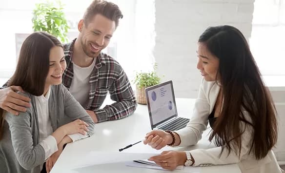 A smiling couple sits at a desk with a professional woman, who is pointing at documents while explaining something. A laptop with charts on the screen is open between them in a bright office setting.
