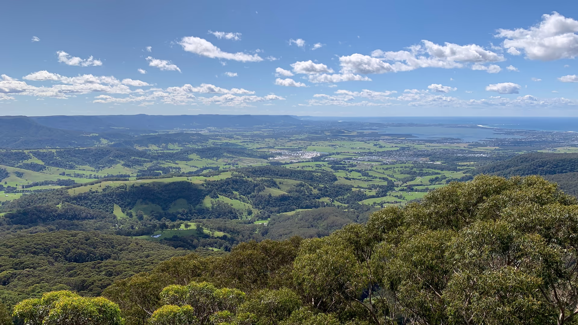 A scenic view from a high vantage point showing rolling green hills, forests, farmland, and distant mountains under a blue sky with scattered clouds. A body of water is visible near the horizon.