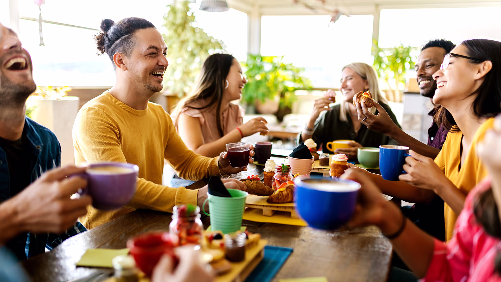 A group of friends sit around a table, laughing and enjoying coffee, pastries, and desserts in a bright, plant-filled café. Cups and plates of food are scattered on the table as everyone smiles and talks.