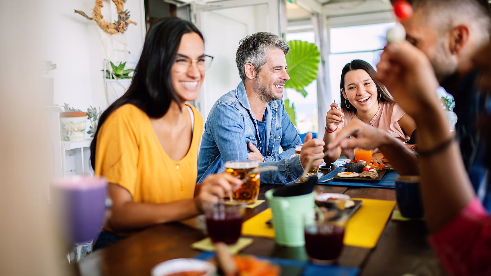 A group of friends sit around a table, laughing and enjoying food and drinks together in a bright, cozy setting.