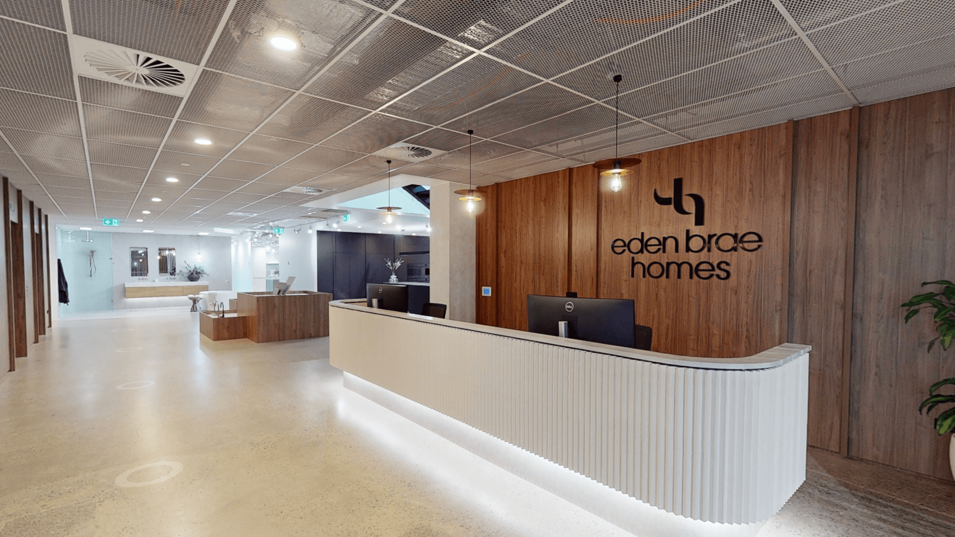 Modern office reception area with a curved white front desk, two computers, wooden accent wall featuring the eden brae homes logo, and bright lighting. Open layout with polished floors and minimalistic decor.