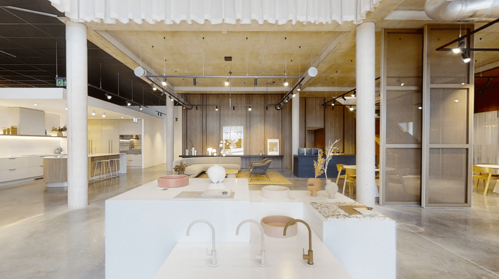 A modern, spacious showroom with minimalist decor, high ceilings, wood and concrete finishes, and displays of designer sinks, faucets, and decorative objects on white platforms.