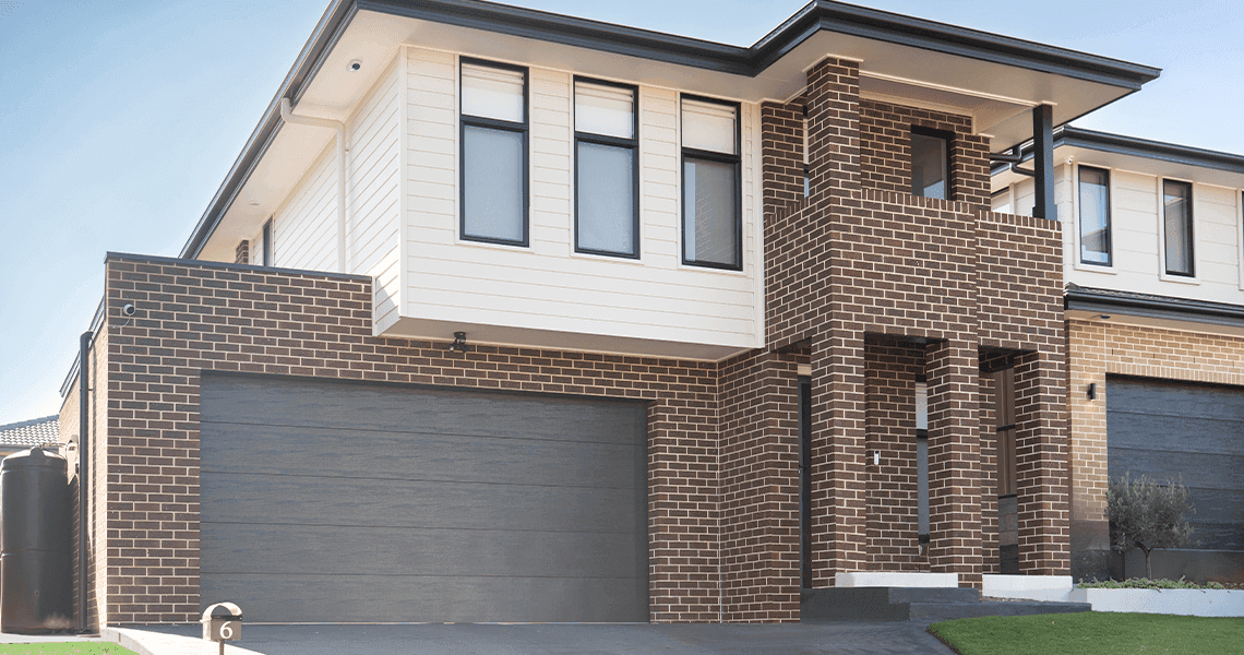 Modern two-story house with brown brick and white panel exterior, large dark gray double garage doors, and multiple rectangular windows on the upper floor. A small covered entrance is at the front.