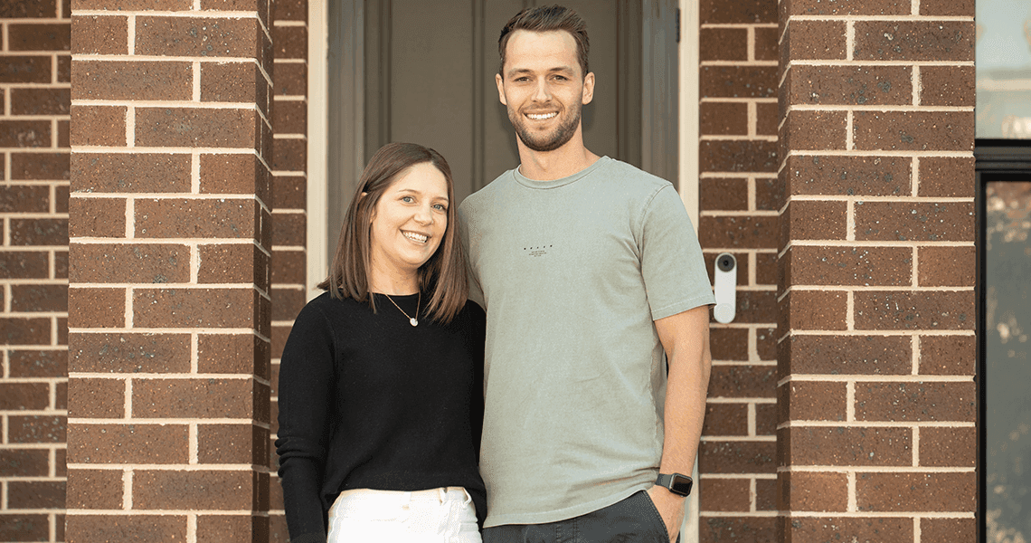 A smiling woman and man stand together in front of a brick house doorway, with the woman wearing a black top and white pants and the man in a light gray t-shirt.