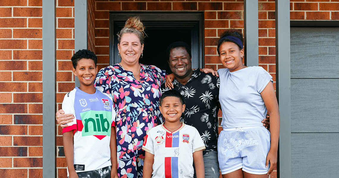 A smiling family of five stands together in front of their brick house. Two adults are in the center, surrounded by three children—two boys and a girl. They are all smiling and dressed casually.