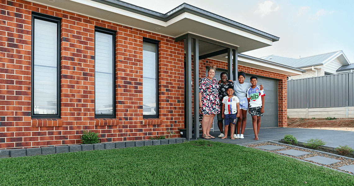 A family of five, including three children and two adults, stands smiling at the entrance of their modern brick house with a neatly maintained lawn and paved walkway in front.
