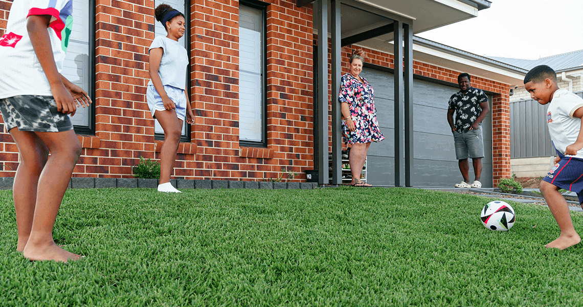 Four children play soccer on a green lawn in front of a brick house, while two adults stand by the entrance watching and smiling. The scene is lively and takes place on a sunny day in a suburban neighborhood.