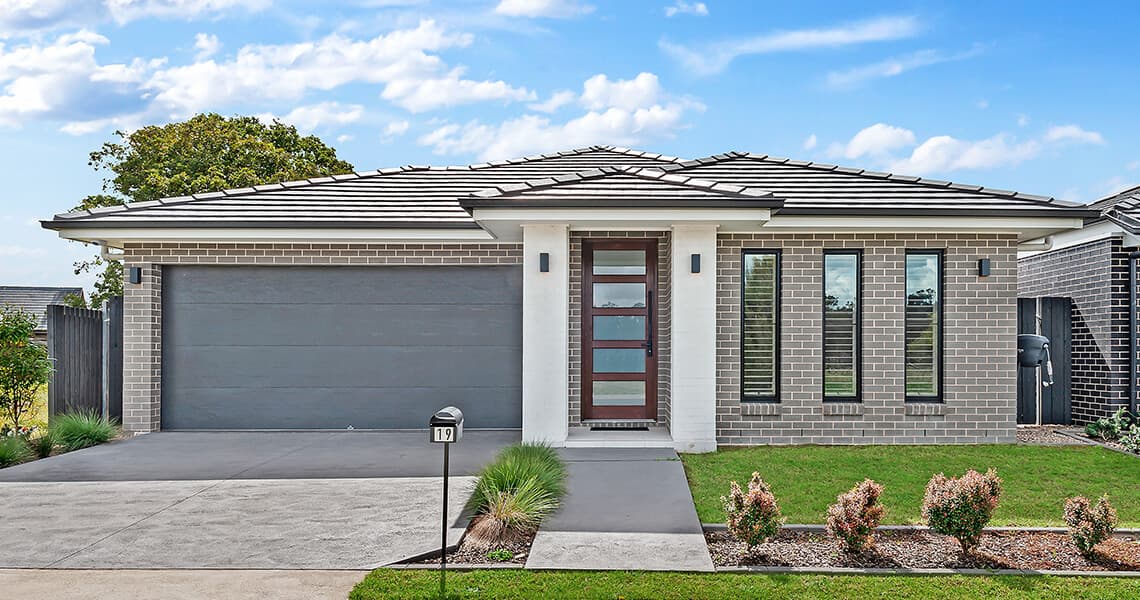 Modern single-story house with gray brick exterior, large double garage, centered glass-panel front door, narrow vertical windows, neat lawn, and small shrubs along a concrete driveway under a blue sky with clouds.