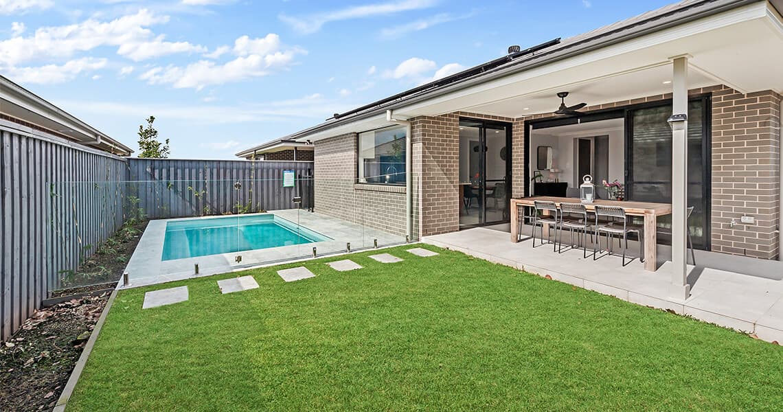 A modern backyard with a rectangular swimming pool, green lawn, stepping stones, and a covered patio area featuring a dining table and chairs next to a brick house.