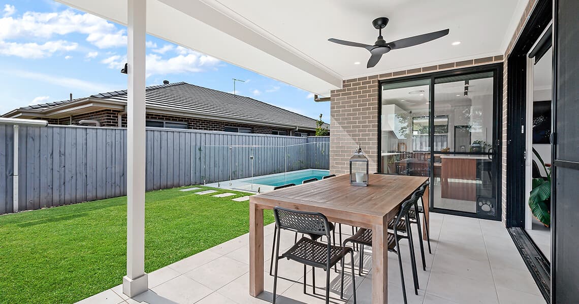 A covered patio with a wooden dining table and six chairs overlooks a backyard with a small swimming pool, green lawn, and a gray wooden fence. Ceiling fan and lantern on the table are visible.