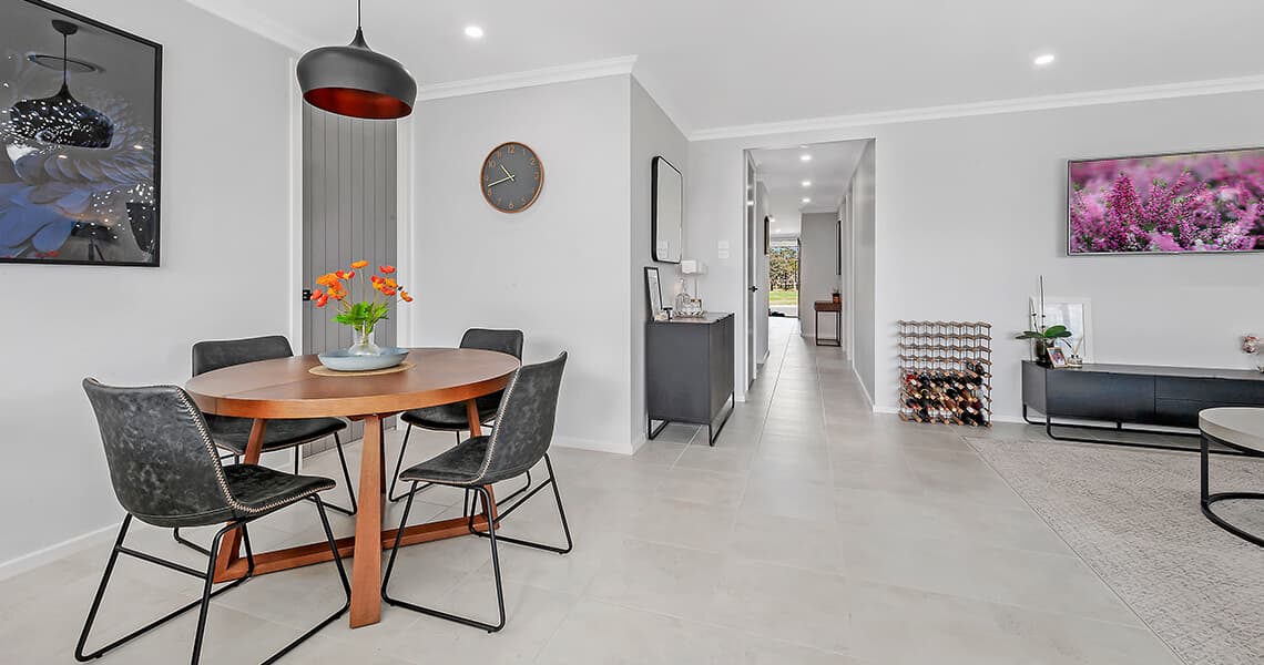 Modern dining and living area with a round wooden table, four black chairs, wall art, a clock, and a TV. The room has light-colored floors and bright lighting, and features minimalistic, contemporary decor.
