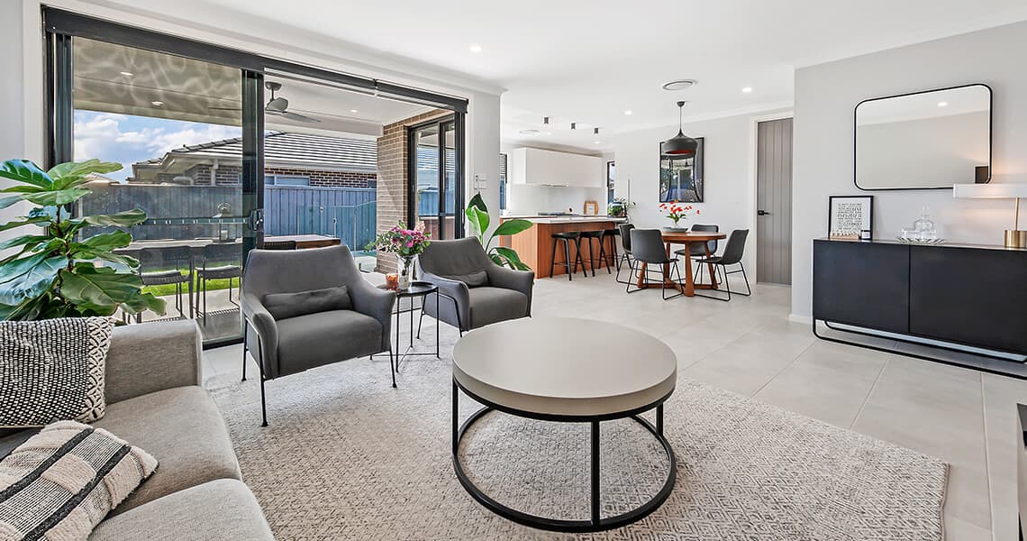 Modern open-plan living room and dining area with neutral tones, stylish furniture, large rug, round coffee table, indoor plants, and sliding glass doors leading to a backyard patio with outdoor seating.