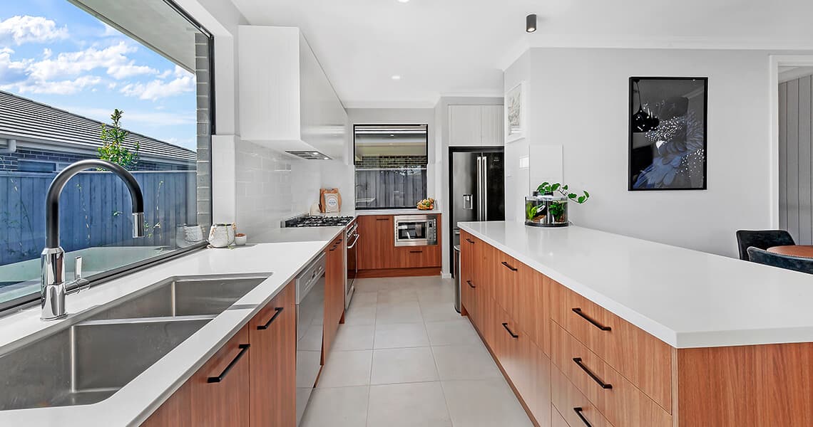 Modern kitchen with large windows, white countertops, stainless steel appliances, wooden cabinets, a double sink, and a kitchen island with plants and decor. Bright natural light fills the space.