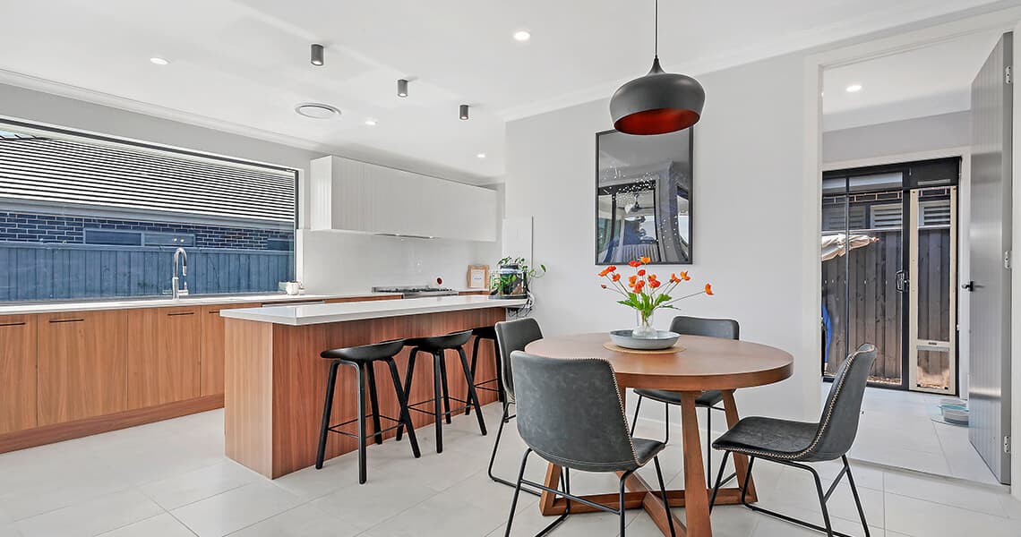 Modern kitchen and dining area with wooden cabinets, a white island with three black barstools, round table with five chairs, vase of orange tulips, and large window letting in natural light.