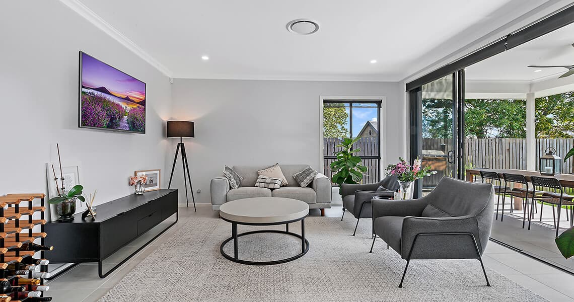Modern living room with a gray sofa, two gray armchairs, round coffee table, TV on the wall, floor lamp, and large glass doors opening to a patio and yard with greenery.