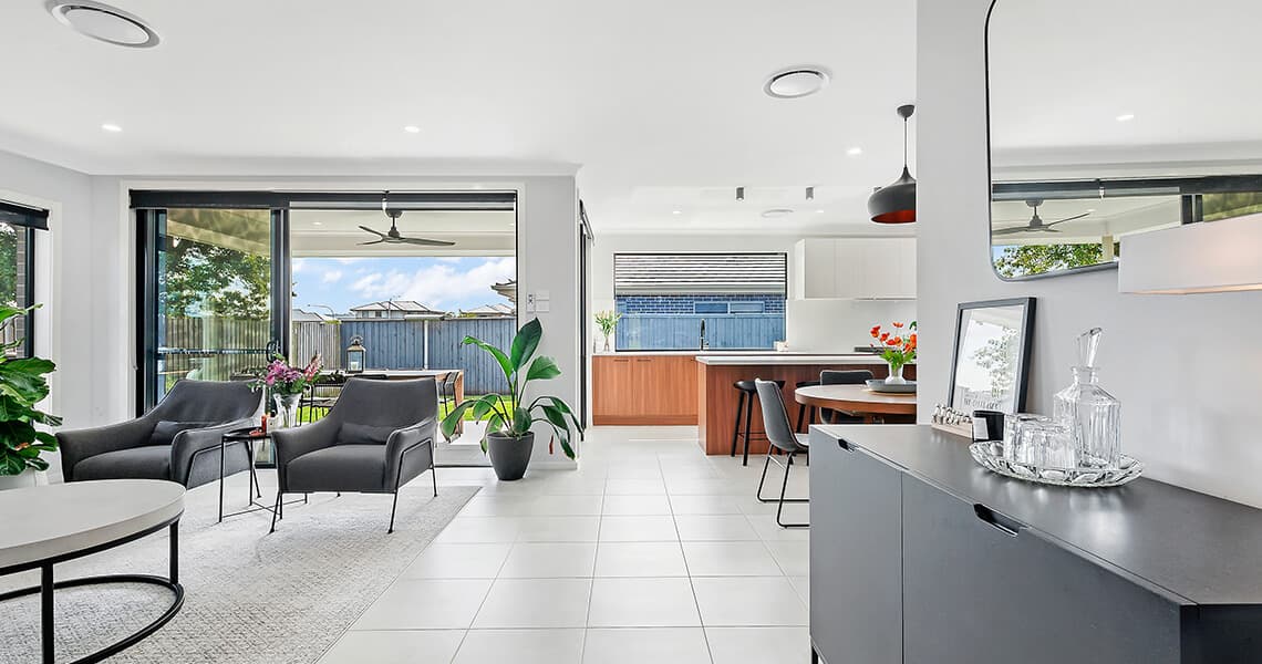 Modern open-plan living and dining area with large windows, neutral décor, gray chairs, indoor plants, a round dining table, and a cabinet with glassware on a tray. Outdoor fenced patio visible through sliding glass doors.